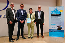 Eine junge Frau in hellgrünem Hosenanzug und drei ältere Herren im Anzug in einem Hörsaal