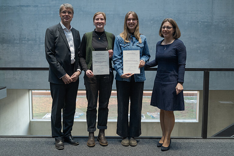 [Translate to English:] Ein Mann und drei Frauen stehen nebeneinander, die beiden jungen Frauen in der Mitte halten eine Urkunde in die Kamera. Alle lächeln.