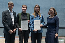 Ein Mann und drei Frauen stehen nebeneinander, die beiden jungen Frauen in der Mitte halten eine Urkunde in die Kamera. Alle lächeln.