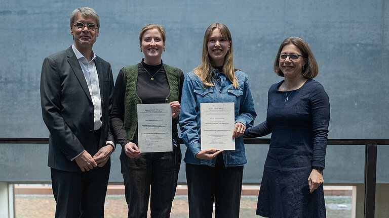 Ein Mann und drei Frauen stehen nebeneinander, die beiden jungen Frauen in der Mitte halten eine Urkunde in die Kamera. Alle lächeln.