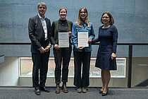 [Translate to English:] Ein Mann und drei Frauen stehen nebeneinander, die beiden jungen Frauen in der Mitte halten eine Urkunde in die Kamera. Alle lächeln.