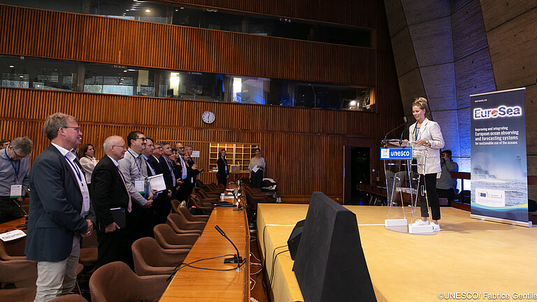 Eine junge Frau hält einen Vortrag auf einer Bühne. Die Menschen im Publikum schauen stehend zu ihr.