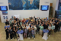 A group of people are standing in a foyer. Some of them point to a screen with the words "Stronger together".