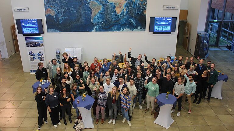 Eine Gruppe Menschen steht in einem Foyer. Einige von ihnen zeigen auf einen Bildschirm mit der Aufschrift "Stronger together".