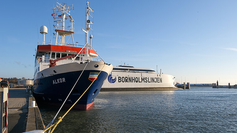 Das Forschungsschiff ALKOR ist im Hafen festgemacht, im Hintergrund ist ein großes Fährschiff zu sehen, auf dem groß der Name "Bornholmslinjen" steht.