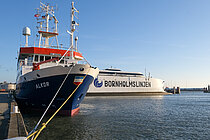 Das Forschungsschiff ALKOR ist im Hafen festgemacht, im Hintergrund ist ein großes Fährschiff zu sehen, auf dem groß der Name "Bornholmslinjen" steht.