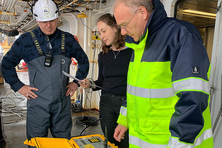 Zwei Männer und eine Frau stehen an Bord eines Schiffes um ein kleines gelbes Steuergerät herum. 