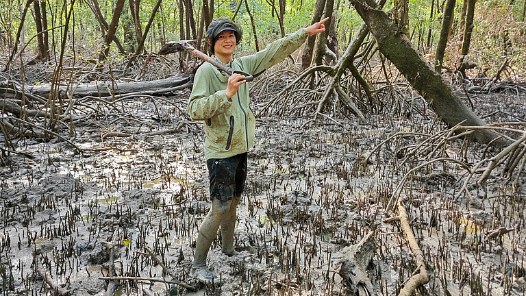 Ein junger Mann mit einer Schaufel über der Schulter zeigt mit dem Finger in die Ferne. Seine Beine sind bis über die Knie mit Schlamm bedeckt.