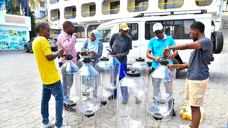 Sechs junge dunkelhäutige Menschen auf einem gepflasterten Hof vor einen weißen Transporter. Sie stehen um zehn große gläserne Gefäße mit schwarzen Deckeln.