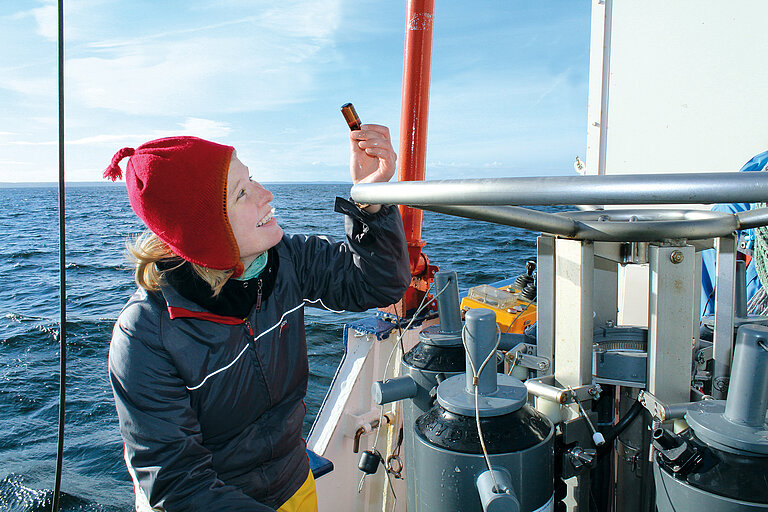 Eine Frau mit roter Mütze auf dem Schiff hält ein braunes Probenfläschchen in die Höhe 