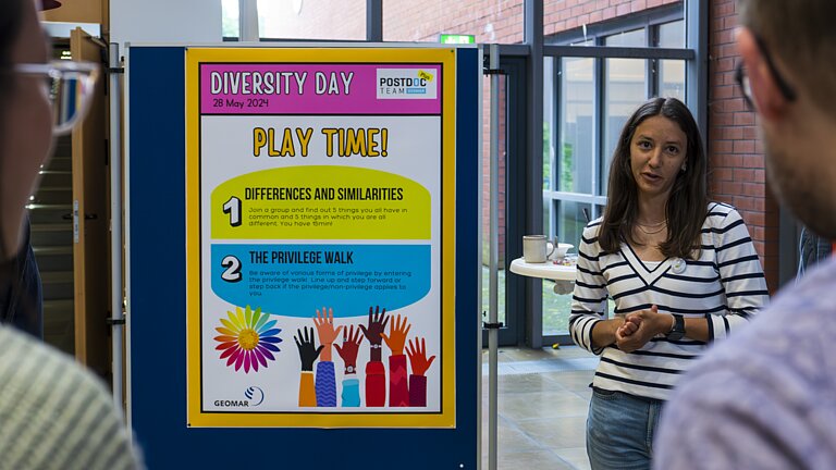 Eine Frau mit langen dunklen Haaren steht rechts neben einer Posterwand. Auf dem Poster sind Spielregeln auf Englisch zu lesen.