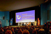 A packed movie theater. A panel discussion is taking place on stage. 