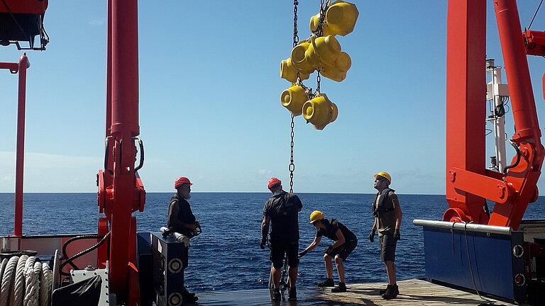 Vier Personen an Bord eines Forschungsschiffes  mit einer Kette, an der einige gelbe kugelförmige Auftriebskörper hängen