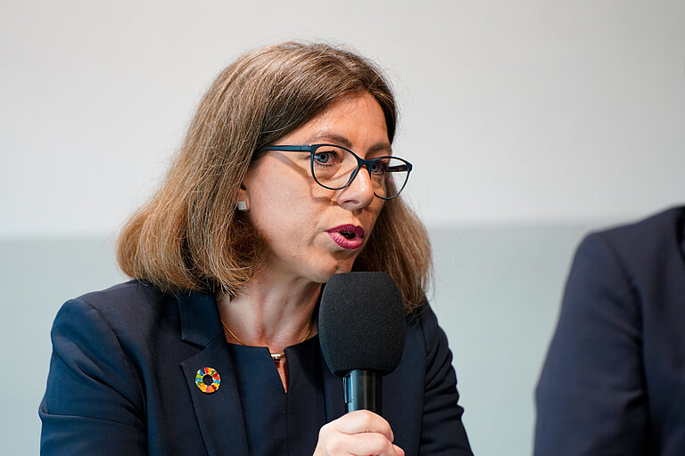Eine Frau mit Brille und halblangen braunen Haaren in einem dunkelblauen Kostüm