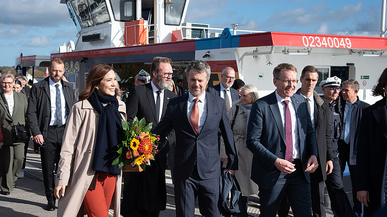 Eine große Gruppe offiziell gekleideter Personen geht an eine Schiff vorbei